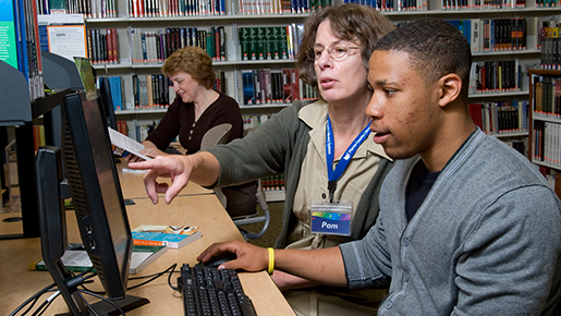 A librarian assists a young man with the computer at a local library while other patrons look on.