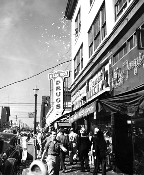 VJ Day crowds in front of Forget-Me-Not Florists and Olberg Drugs at Pacific and 4th on August 14, 1945. Confetti flutters from the third floor windows. Puget Sound Naval Shipyard