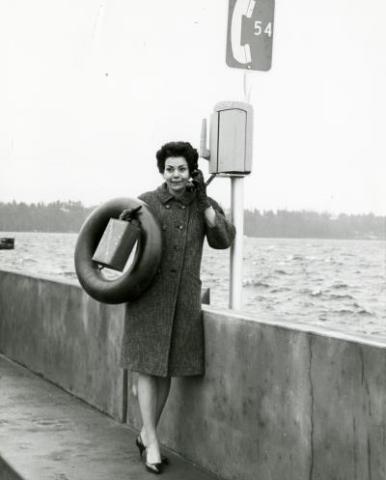 Black and white photograph of a woman using an emergency telephone and holding a tire and gasoline can on the Evergreen Point Floating Bridge.