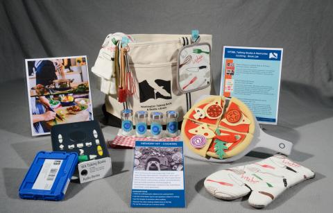 A large bag surrounded by an oven mitt, potholder and dish towel, cooking utensils, jars, a felt pizza set, a book player and cartridge, and instructional cards