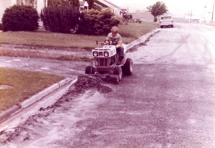 Photo of a person cleaning up the ash on a residential street using a small bulldozer. 