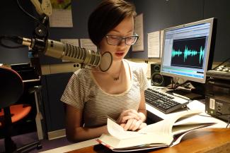 Woman sitting at a desk with a book open and a microphone in front of her. In the background is a computer display with audio software on the screen. 