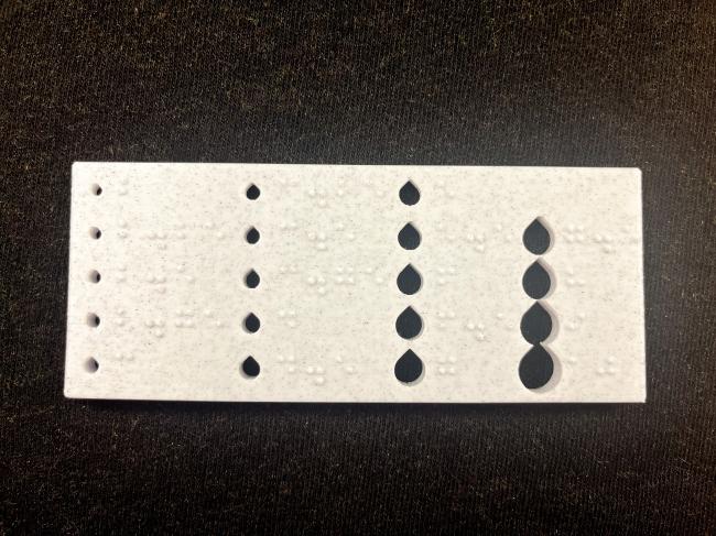Braille knitting needle gauge, which is a horizontal rectangle with holes for knitting needles that get progressively larger