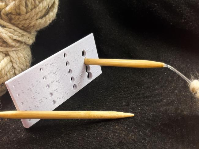 A knitting needle being sized with the braille knitting needle gauge.