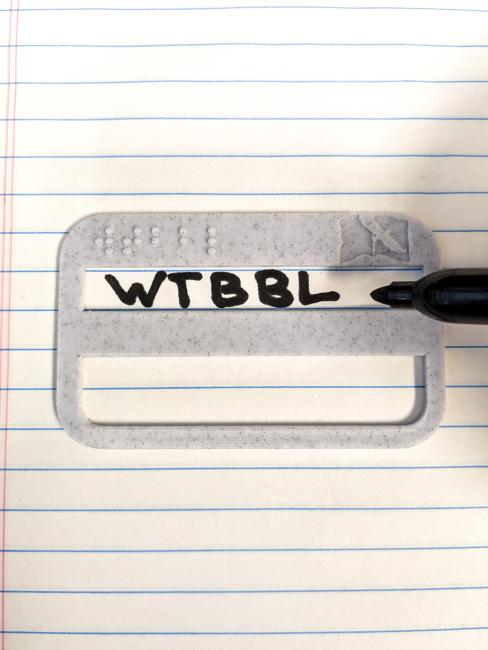 Signature guide with "wtbbl" in braille and the WTBBL logo on it. The smaller box has been used to write "WTBBL" on notebook paper.